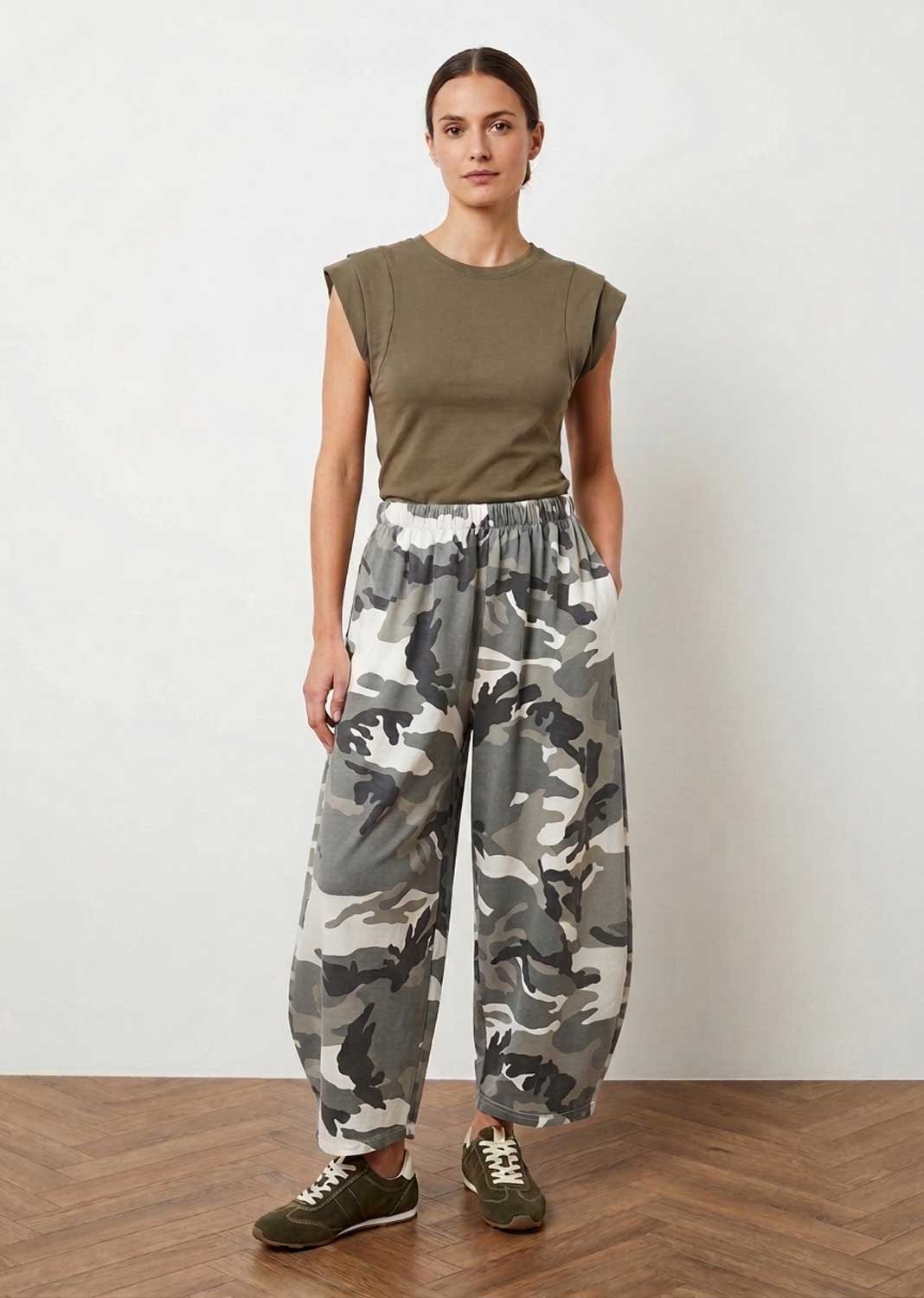 A woman stands in a studio wearing a sleeveless olive green top and loose camouflage print pants in shades of gray, white, and green, paired with olive green sneakers.