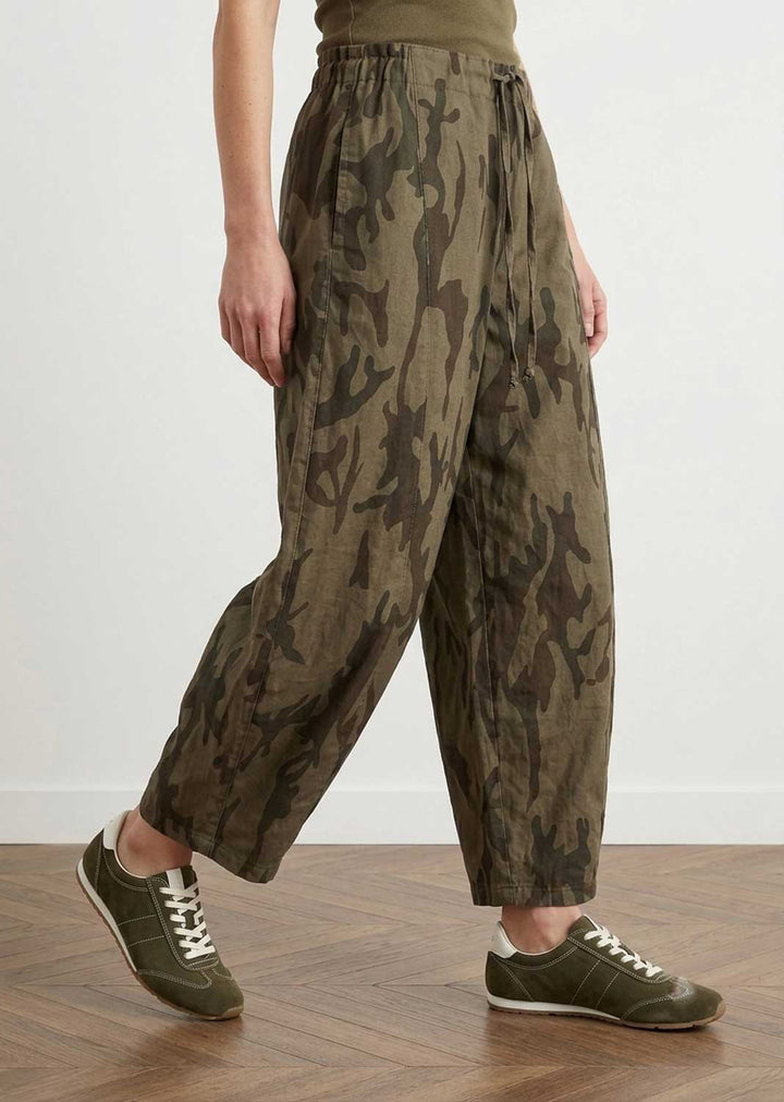 A person wearing loose, wide-leg camouflage trousers in olive green and brown, paired with olive sneakers, standing on a wooden floor against a white wall.