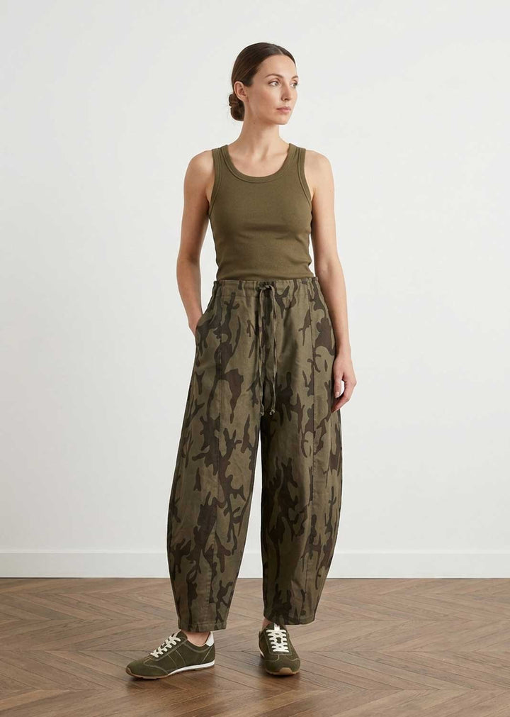 A woman stands in a studio wearing an olive green ribbed tank top and olive green wide-leg pants with a darker abstract pattern, paired with olive green sneakers, against a white wall and wood floor.