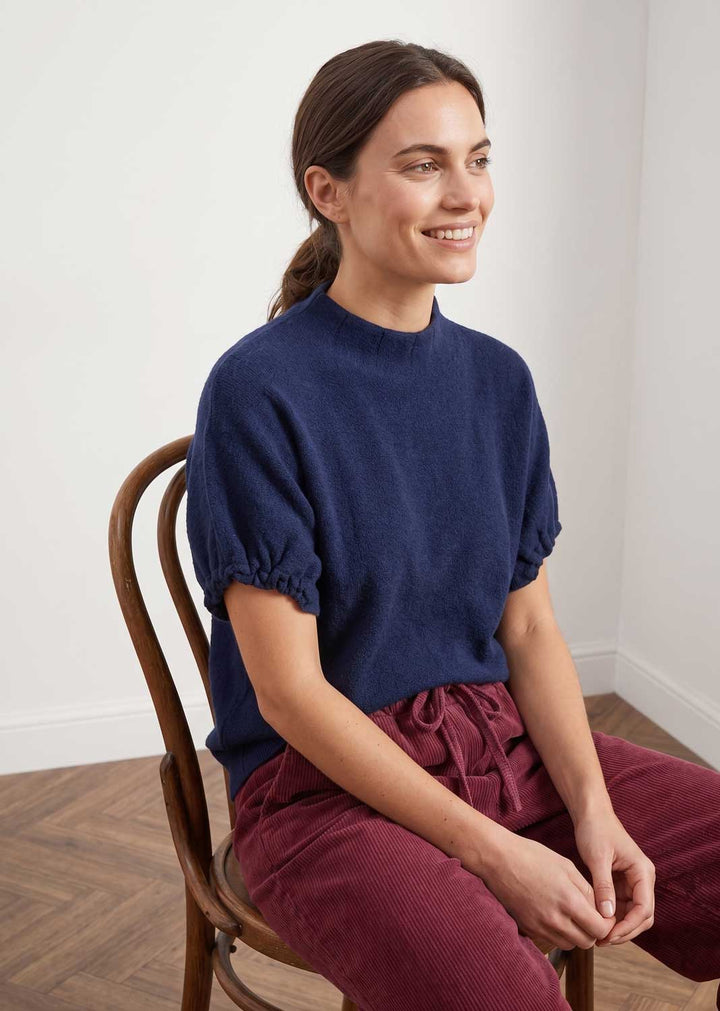 A woman sitting on a wooden chair wearing a navy blue short-sleeve mock neck knit top and burgundy corduroy pants in a bright room with white walls and a wood floor.