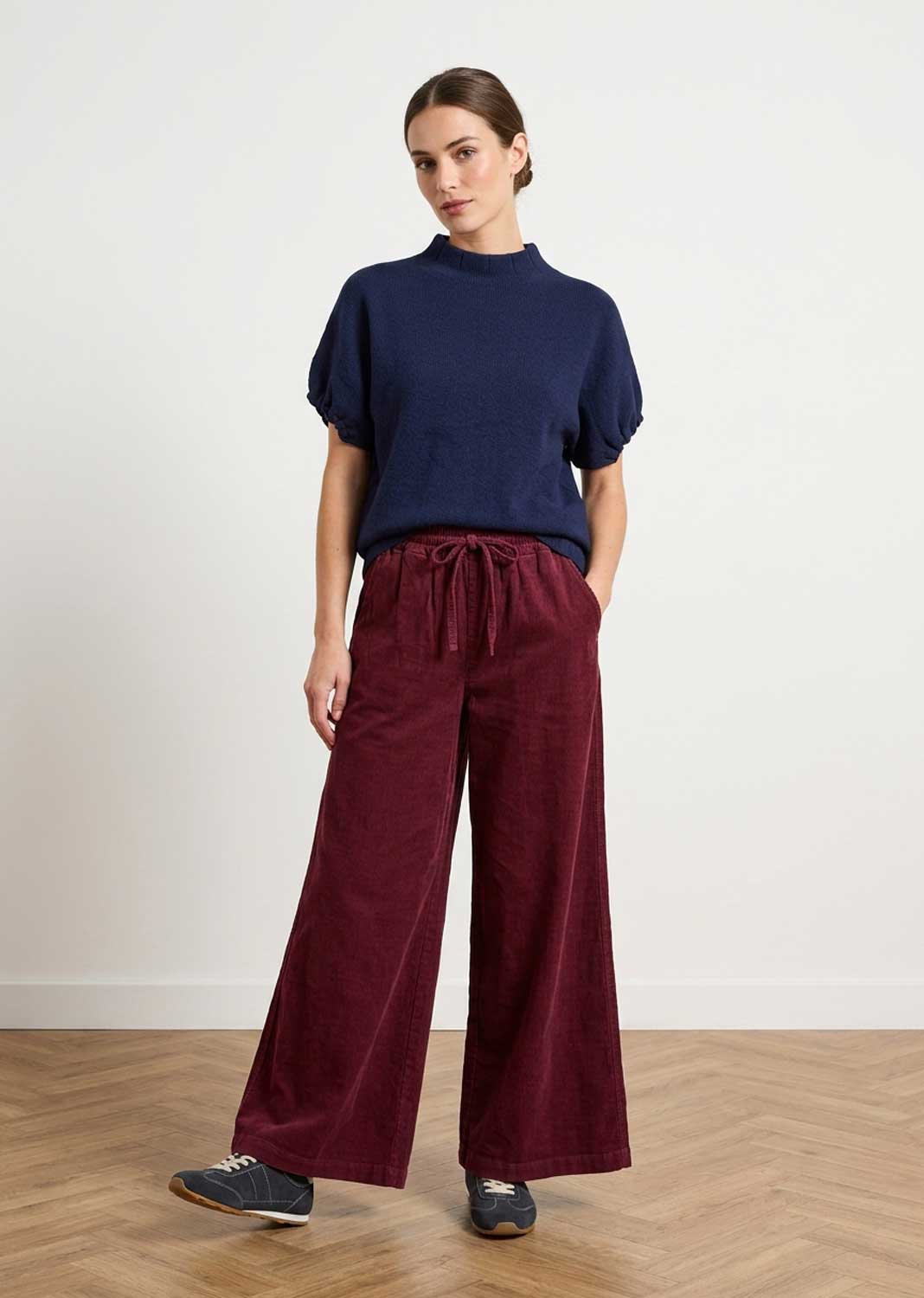 A woman modeling a navy blue short-sleeve knit top and burgundy wide-leg corduroy pants with a drawstring waist, standing on a light wood floor against a white wall.