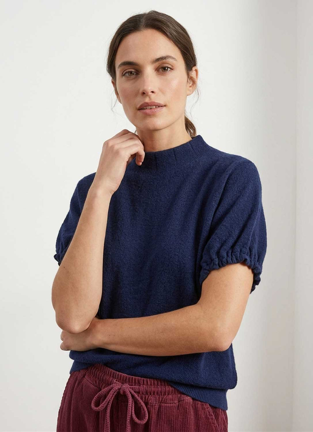 A woman wearing a navy blue short-sleeve knit top with gathered puff sleeves and burgundy corduroy pants, standing against a plain light gray background.