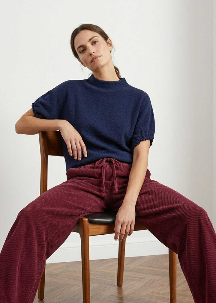 A woman sits on a wooden chair wearing a navy blue short-sleeve knit top and burgundy drawstring trousers in a minimalist room with a white wall and wood floor.