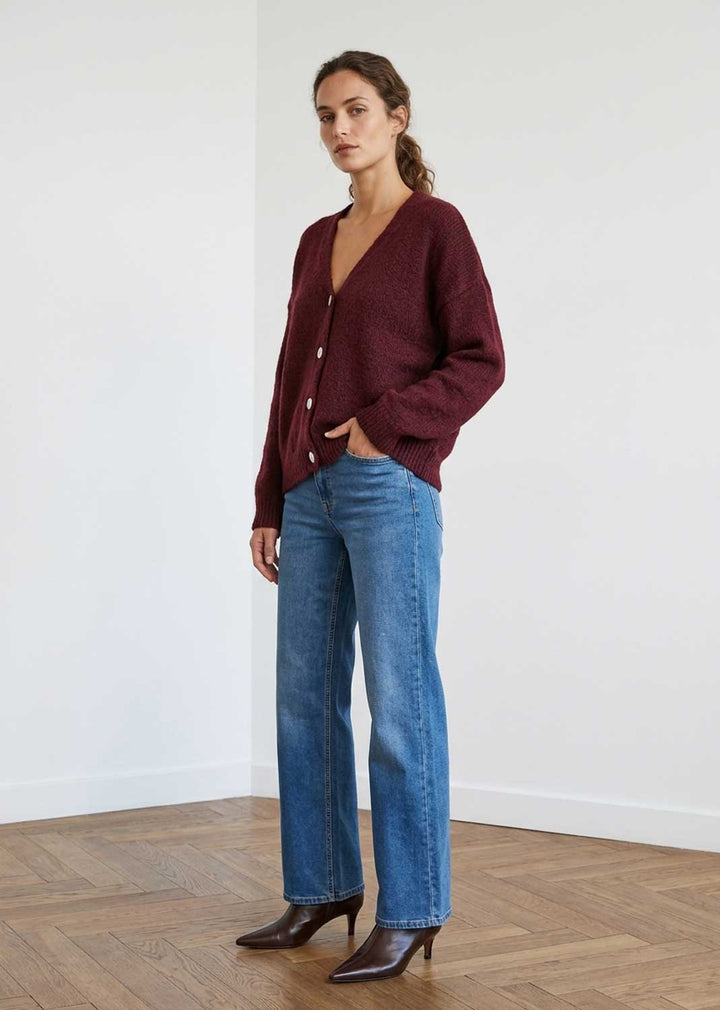 A woman stands in a minimalist room with white walls and a wood floor, wearing a maroon button-up cardigan, blue jeans, and dark brown pointed-toe ankle boots.