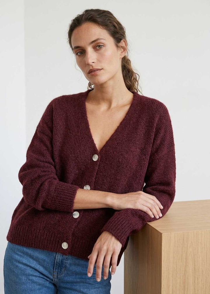A woman wearing a maroon knit cardigan with mother of pearl buttons and blue jeans, leaning against a light wood block in a minimalist indoor setting.