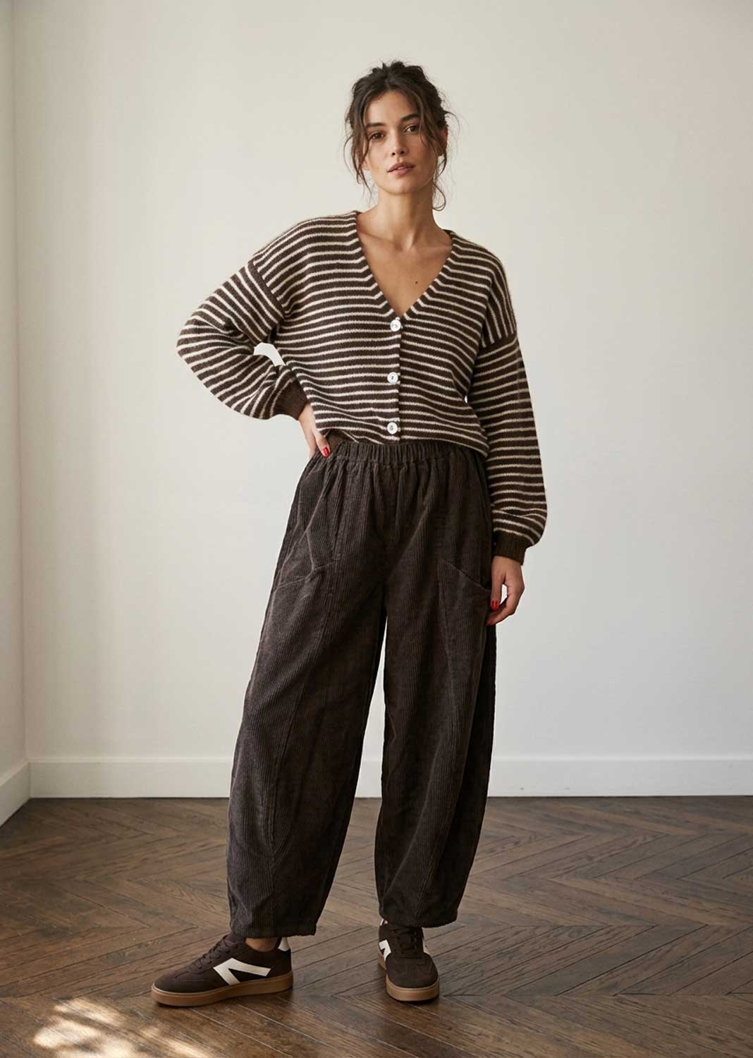 A woman stands in a minimalist room with a white wall and wood floor, wearing a brown and beige horizontally striped long-sleeve button-up cardigan, dark brown wide-leg corduroy pants, and brown sneakers.