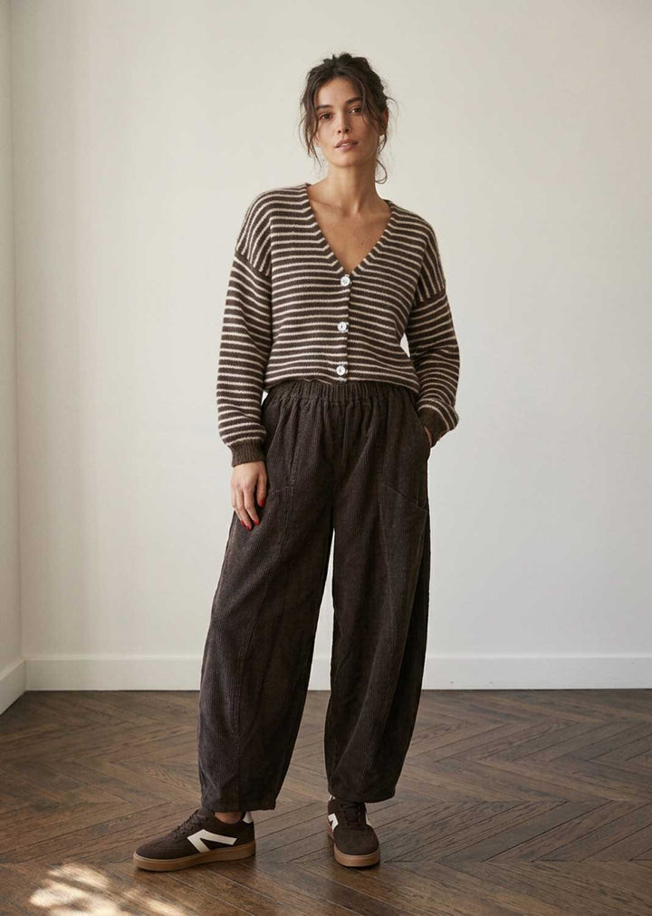 A woman stands in a minimalist room with a white wall and wood floor, wearing a brown and beige striped button-up cardigan, dark brown wide-leg trousers, and brown sneakers.
