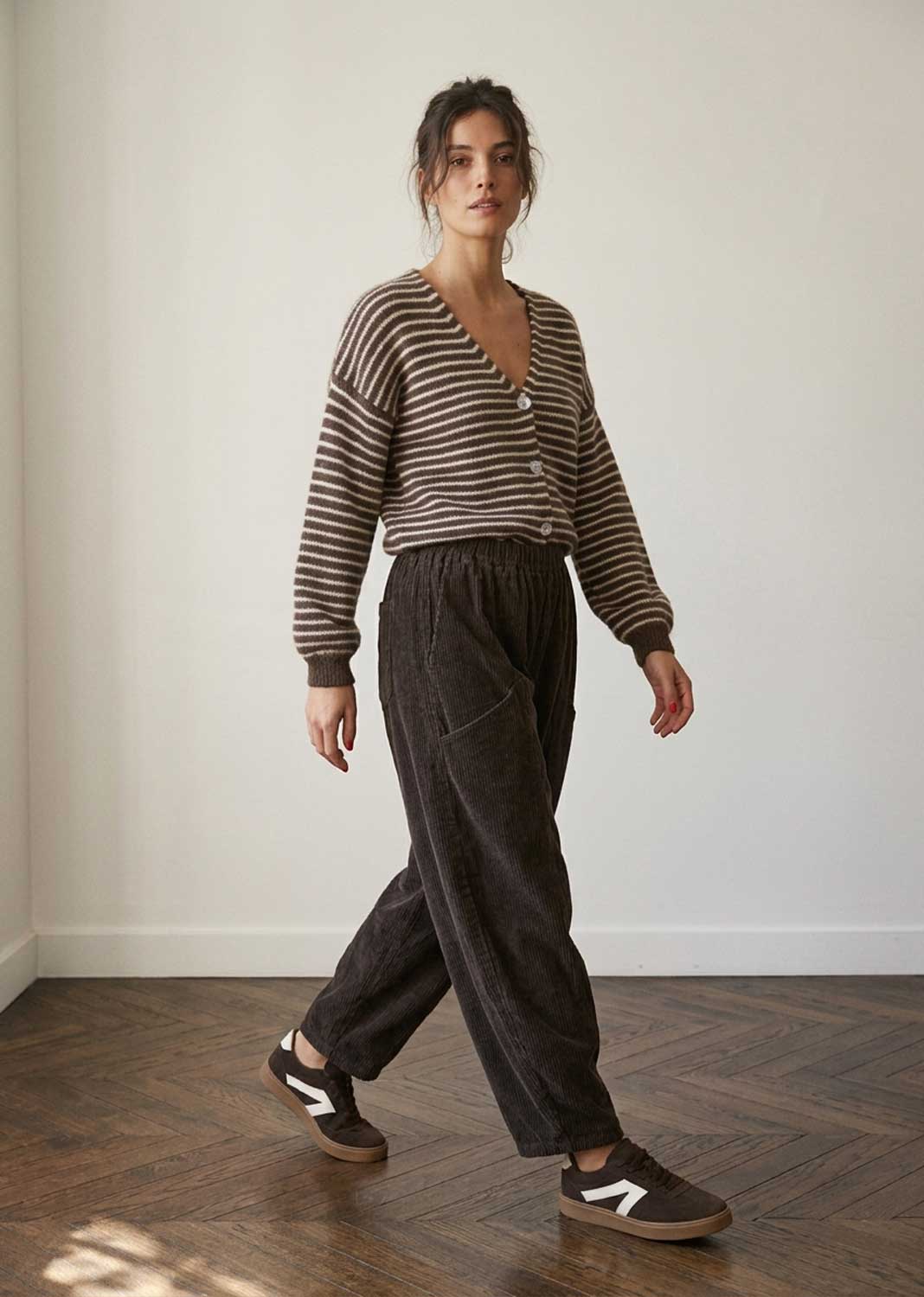 A woman modeling a brown and beige striped knit cardigan, dark charcoal corduroy trousers, and brown suede sneakers with white side stripes, standing in a minimalist room with a white wall and wooden parquet floor.