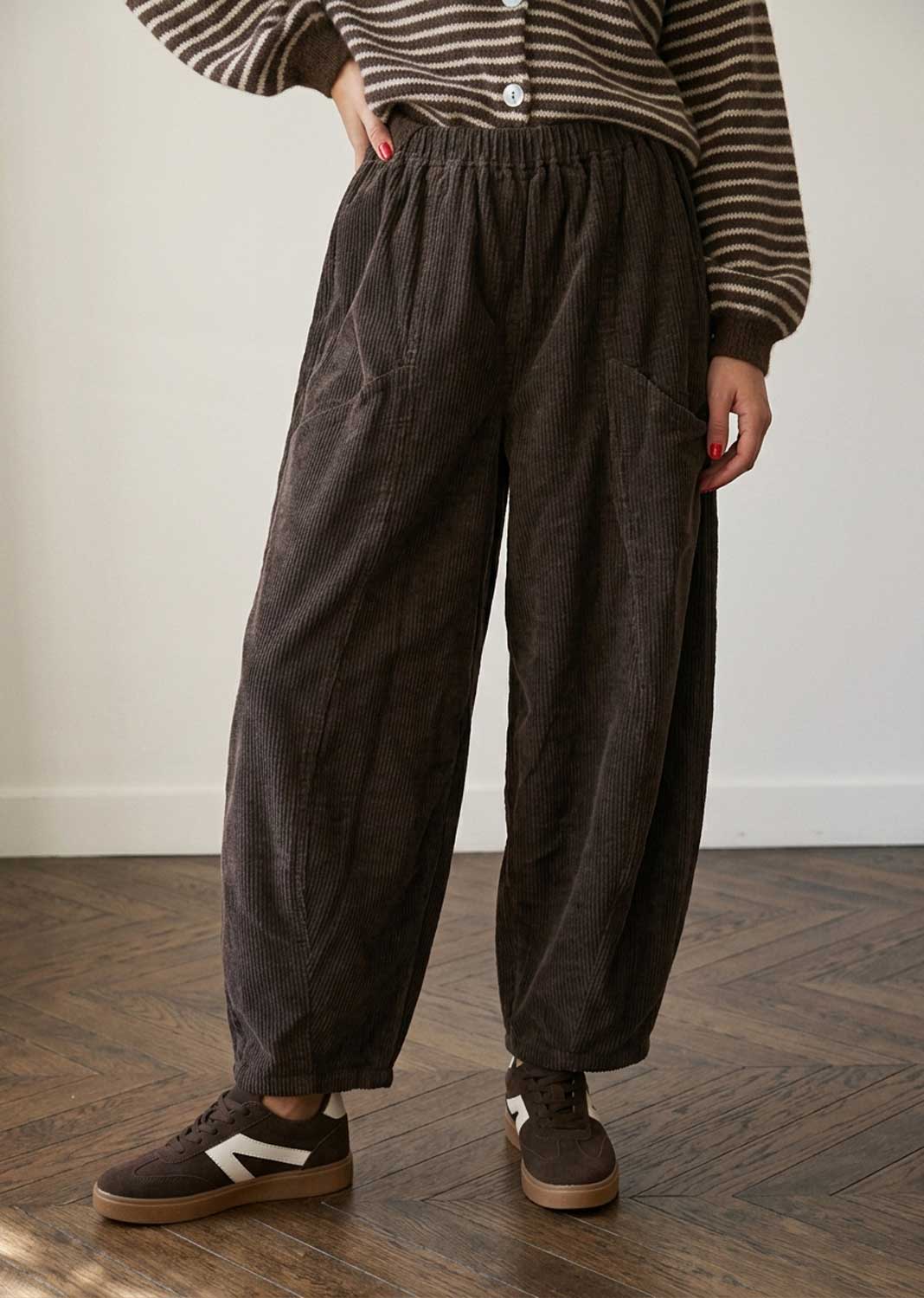 A person wearing loose dark brown corduroy pants with an elastic waistband, paired with a striped long-sleeve top and brown sneakers, standing on a wooden floor against a plain wall.