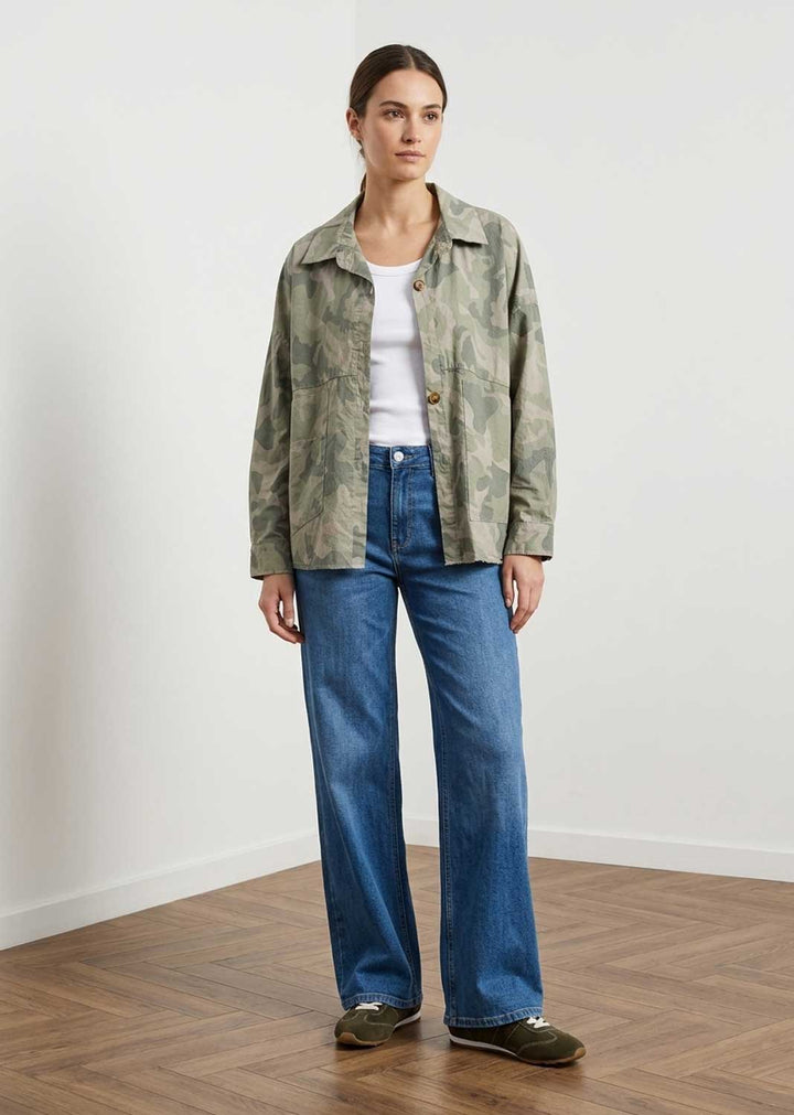 A woman stands in a minimalist room with a white wall and wood floor, wearing a green camouflage jacket over a white top and blue wide-leg jeans.