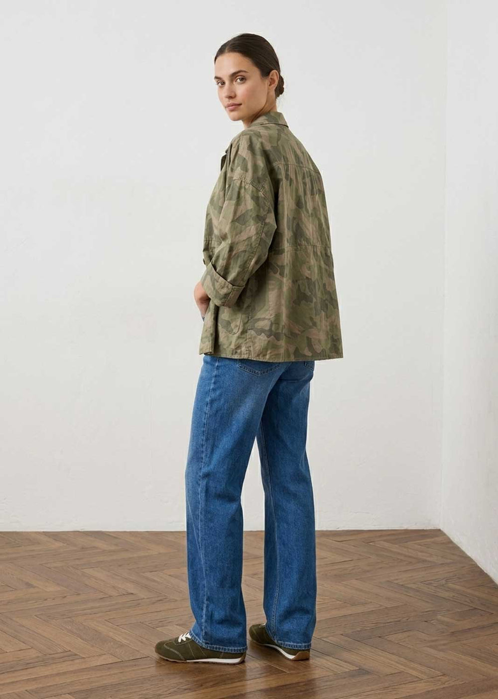 A woman stands in profile wearing a loose olive-green camouflage jacket, blue jeans, and green sneakers in a minimalist room with a white wall and wood herringbone floor.