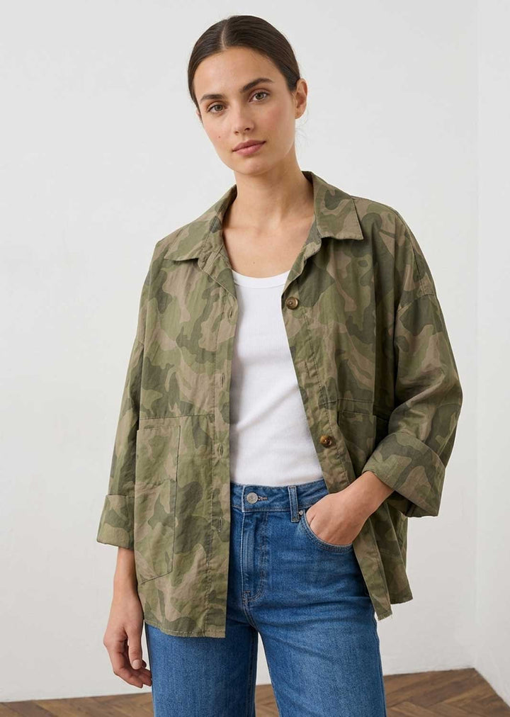 A woman wearing a green camouflage button-up shirt over a white tank top and blue jeans, standing against a plain white wall.