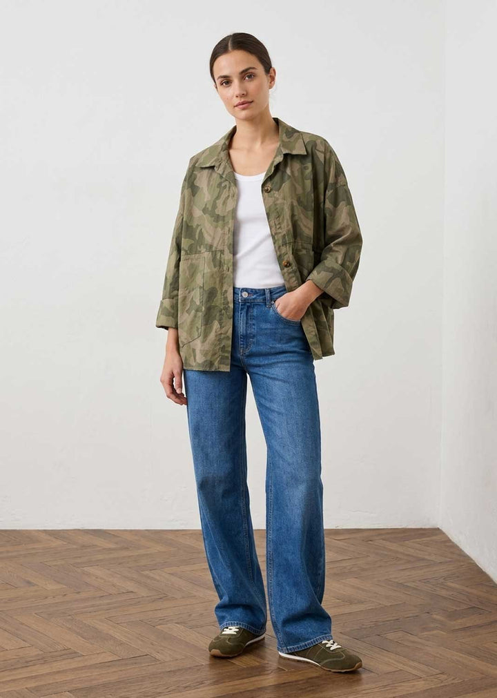 A woman stands in a studio wearing a green camouflage button-up shirt over a white t-shirt, blue jeans, and olive green sneakers.