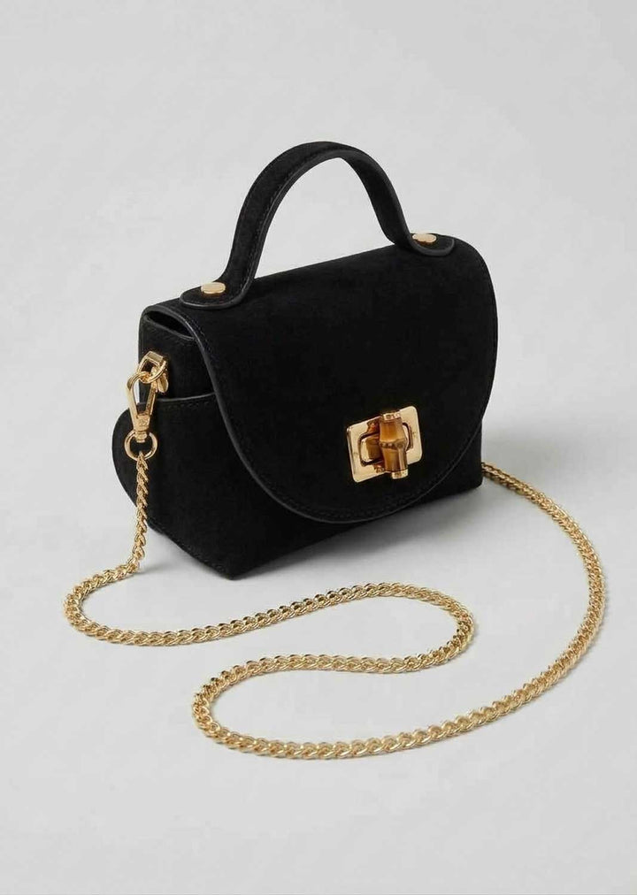 A black suede top-handle handbag with a gold-tone chain strap and a gold clasp, displayed on a light gray background.