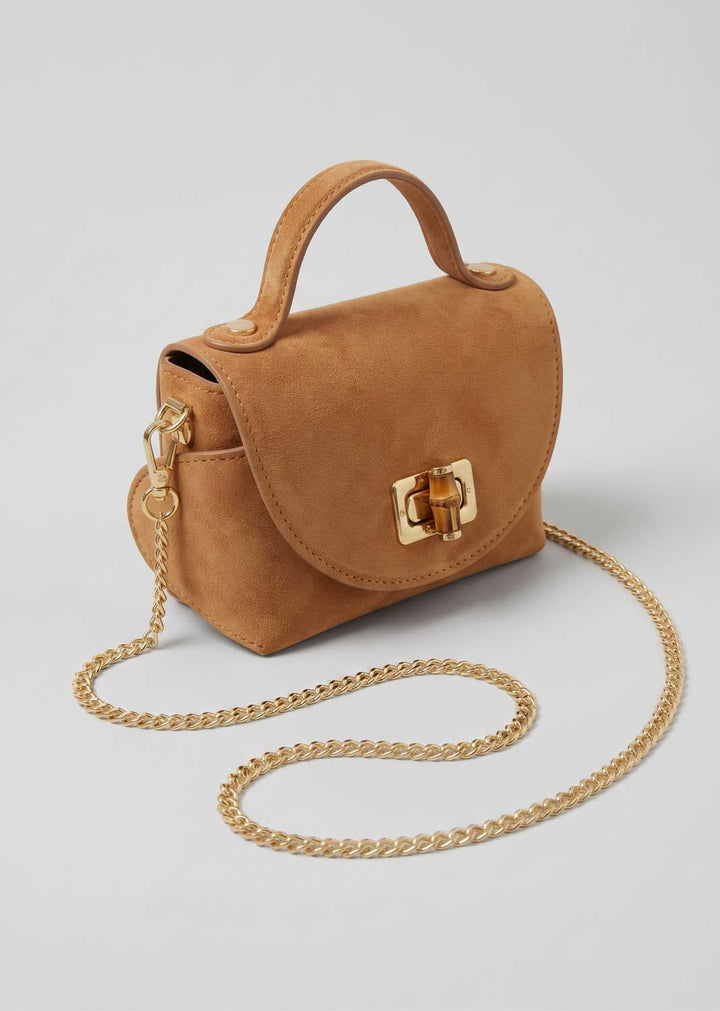 A small tan suede handbag with a top handle, gold-tone turn-lock clasp, and a gold chain shoulder strap, photographed on a light gray background.