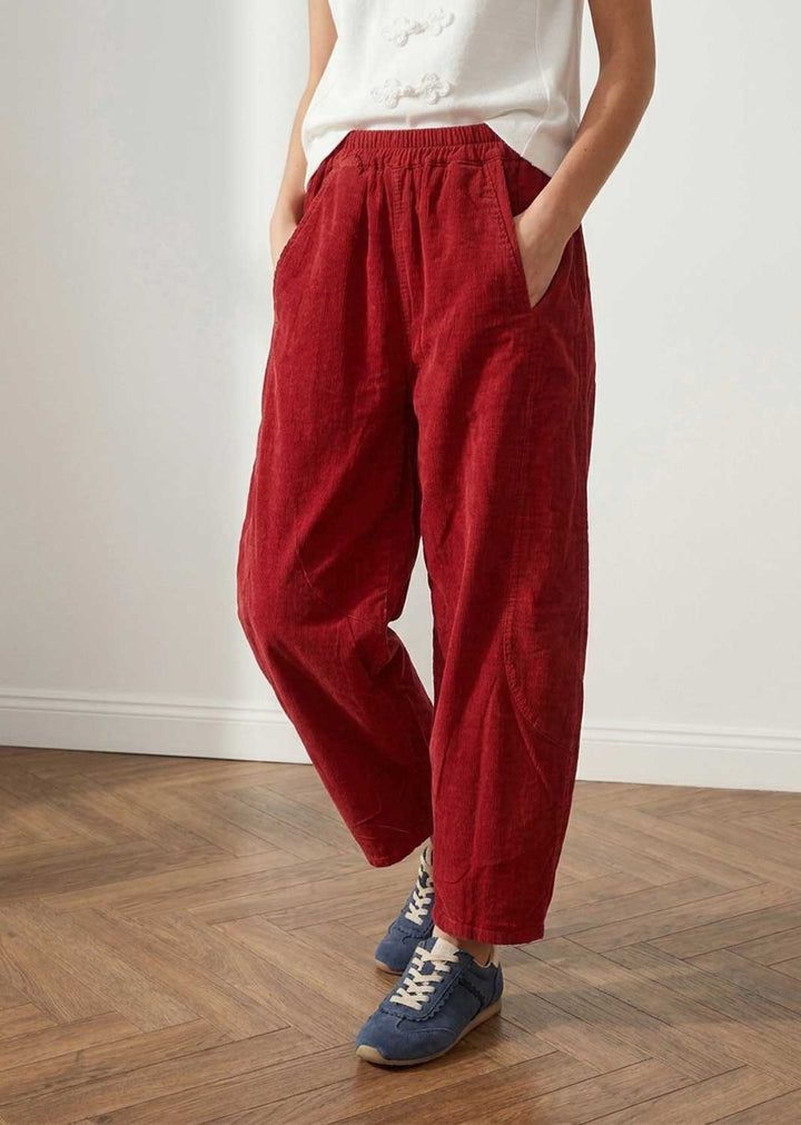 A person wearing loose, barrel leg red corduroy pants with an elastic waistband and side pockets, paired with a white top and blue sneakers, standing on a wooden floor against a white wall.