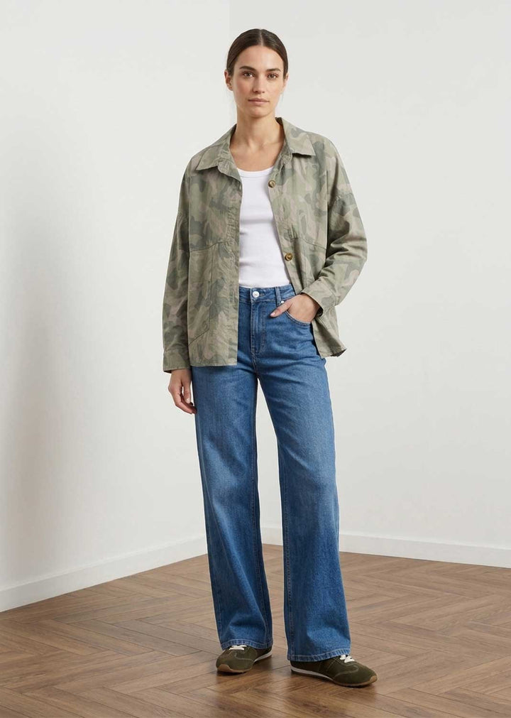A woman stands in a minimalist room with a white wall and wood floor, wearing a long-sleeve button-up shirt jacket with a green and beige abstract pattern, a white t-shirt, wide-leg blue jeans, and dark green slip-on shoes.
