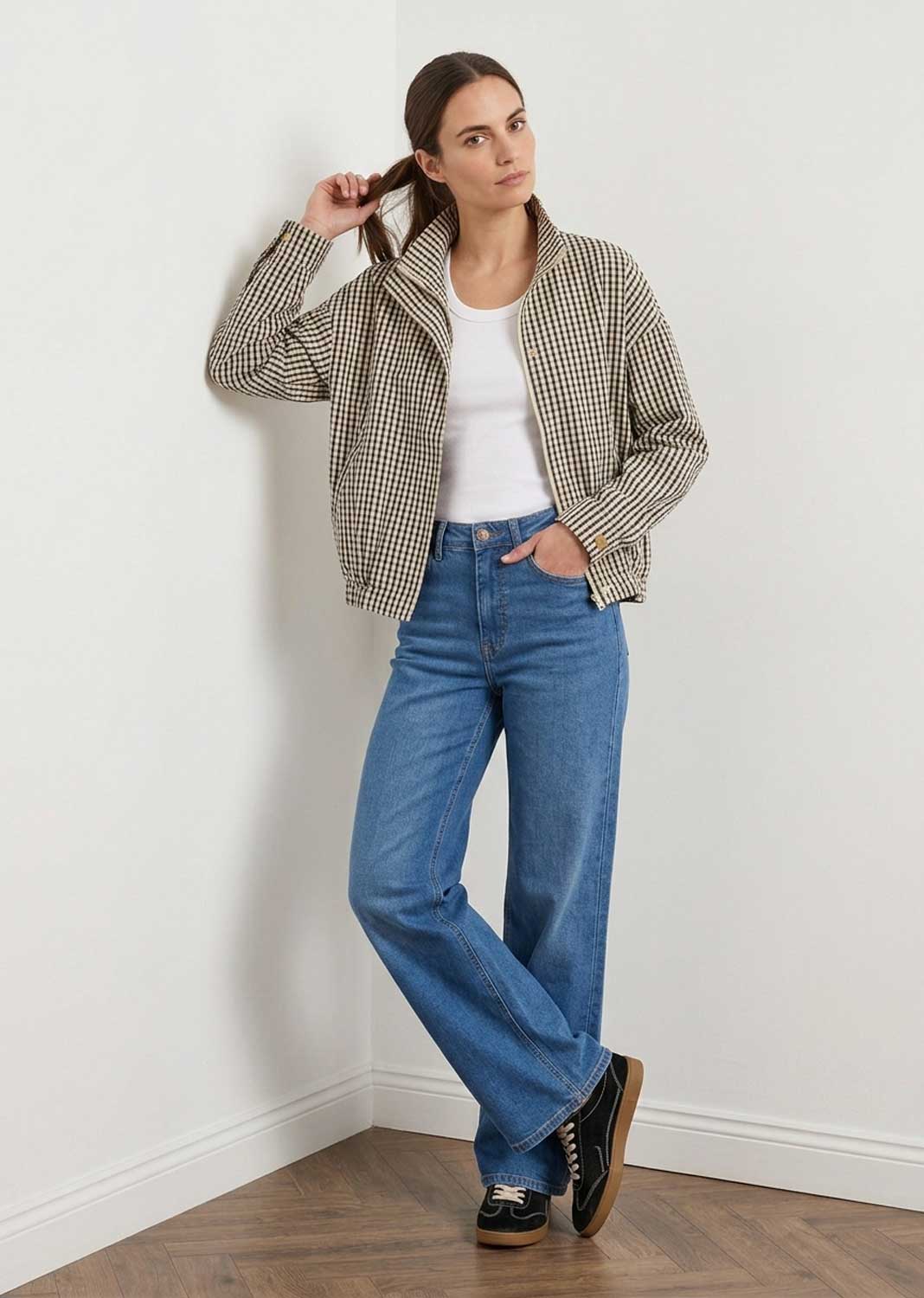 A woman stands in a white room corner wearing a beige and black gingham check windbreaker bomber jacket  over a white top, blue jeans, and black loafers.