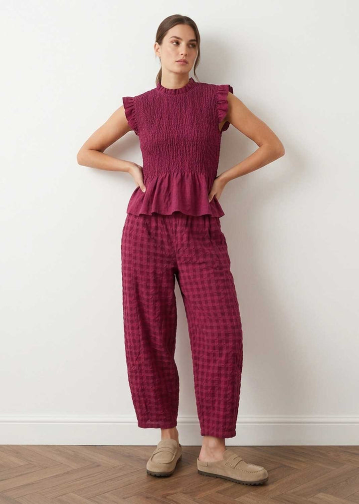 A woman stands against a white wall wearing a matching two-piece outfit with a smocked peplum top and wide-leg plaid pants in a deep magenta color, paired with beige slip-on shoes.
