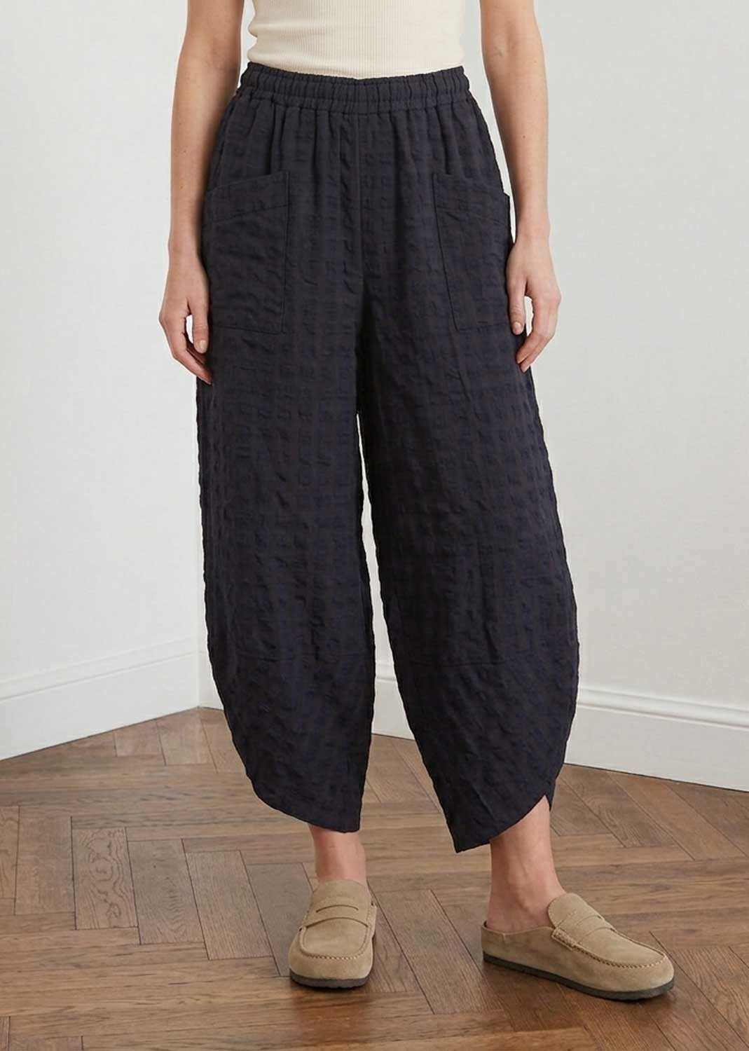 A person wearing loose, wide-leg cropped pants in a dark navy textured fabric, paired with a cream top and beige slip-on loafers, standing on a wooden floor against a white wall.