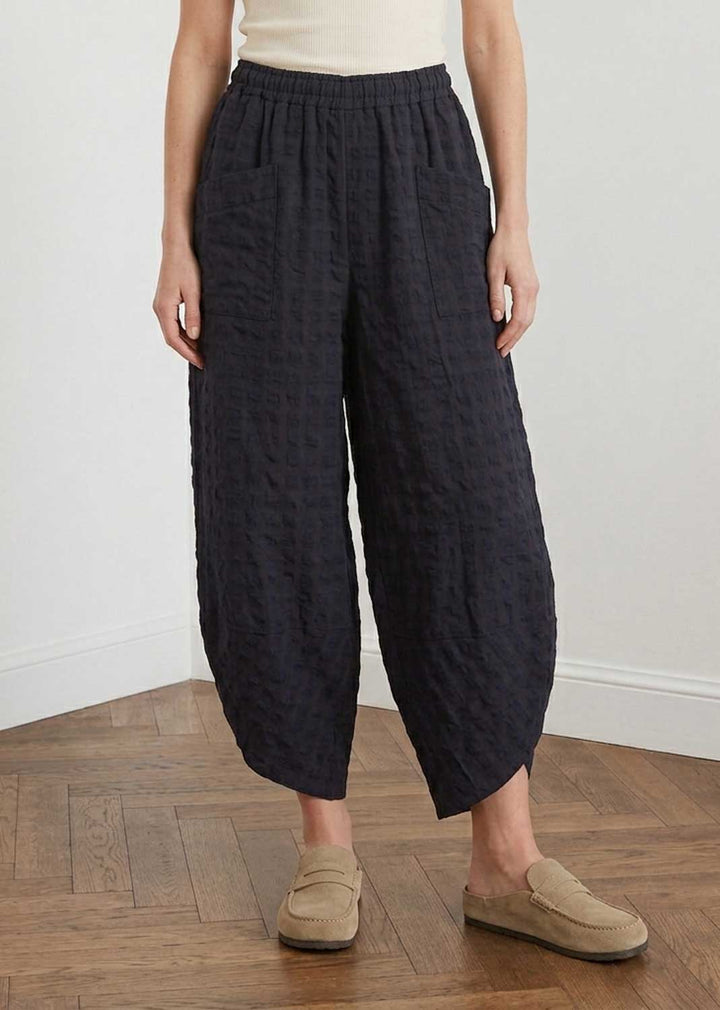 A person wearing loose, wide-leg cropped pants in a dark navy textured fabric, paired with a cream top and beige slip-on loafers, standing on a wooden floor against a white wall.