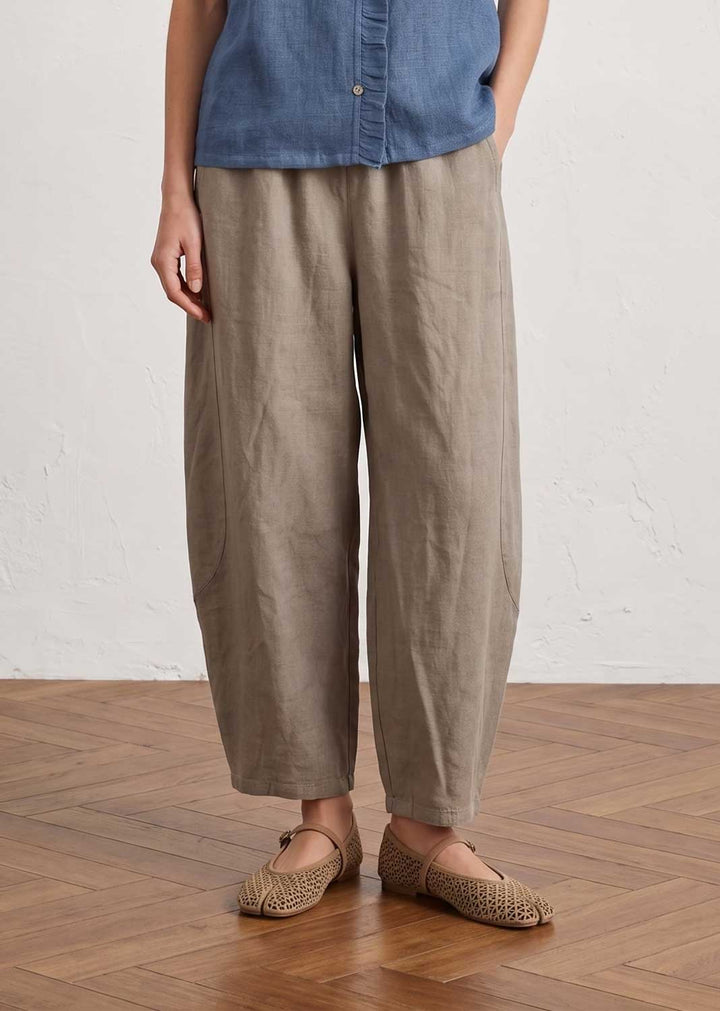 A person wearing loose, wide-leg taupe linen trousers, a blue denim shirt, and woven flat shoes, standing on a wooden parquet floor against a white textured wall.