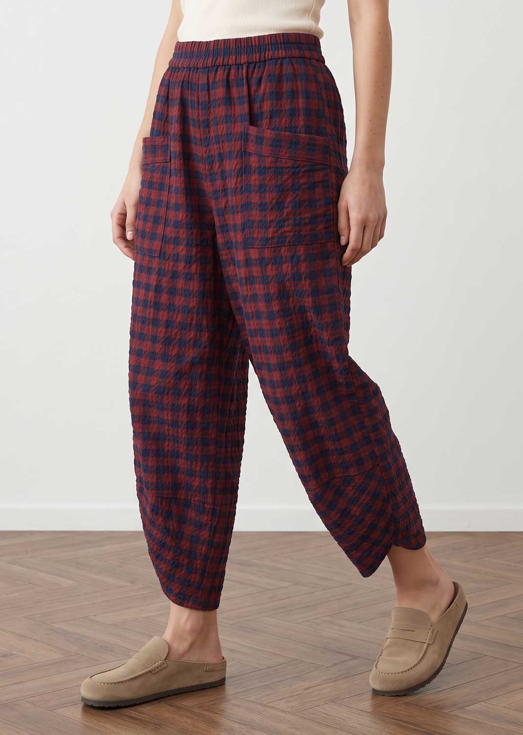 A person wearing loose, gathered plaid pants in dark red and navy, paired with beige slip-on loafers, standing on a wood floor against a white wall.