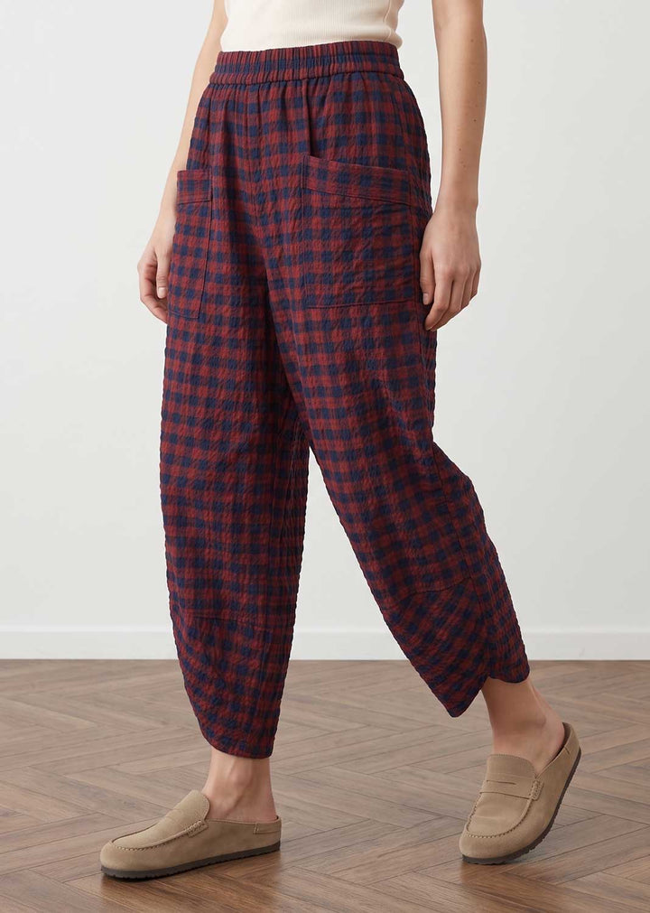 A person wearing loose, gathered plaid pants in dark red and navy, paired with beige slip-on loafers, standing on a wood floor against a white wall.
