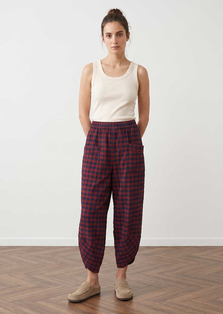 A woman stands in a studio wearing loose red and navy plaid cropped pants with an elastic waist, paired with a cream sleeveless top and beige slip-on shoes.