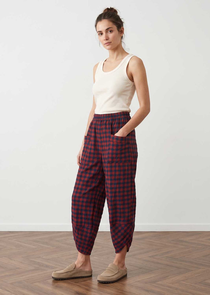 A woman modeling loose, tapered trousers in a red and navy blue plaid pattern, paired with a cream tank top and beige slip-on shoes, standing in a minimalist room with a white wall and wooden floor.