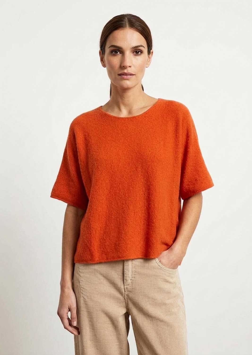 A woman modeling a short-sleeve, crew-neck knit top in a vibrant orange color, paired with beige trousers, against a plain light gray background.