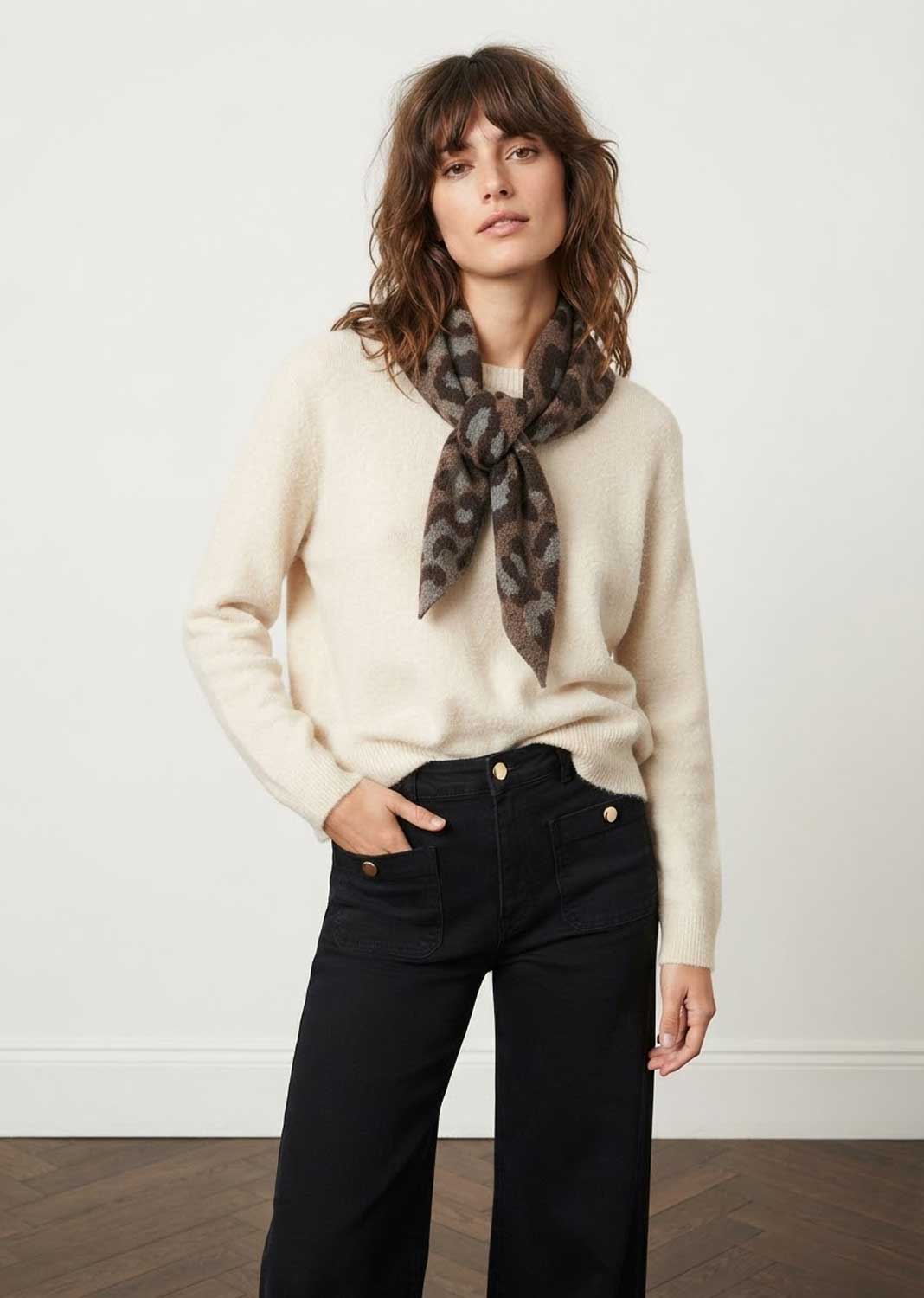 A woman modeling a cream knit sweater and black wide-leg jeans, accessorized with a brown and black patterned scarf, standing against a plain off-white wall.