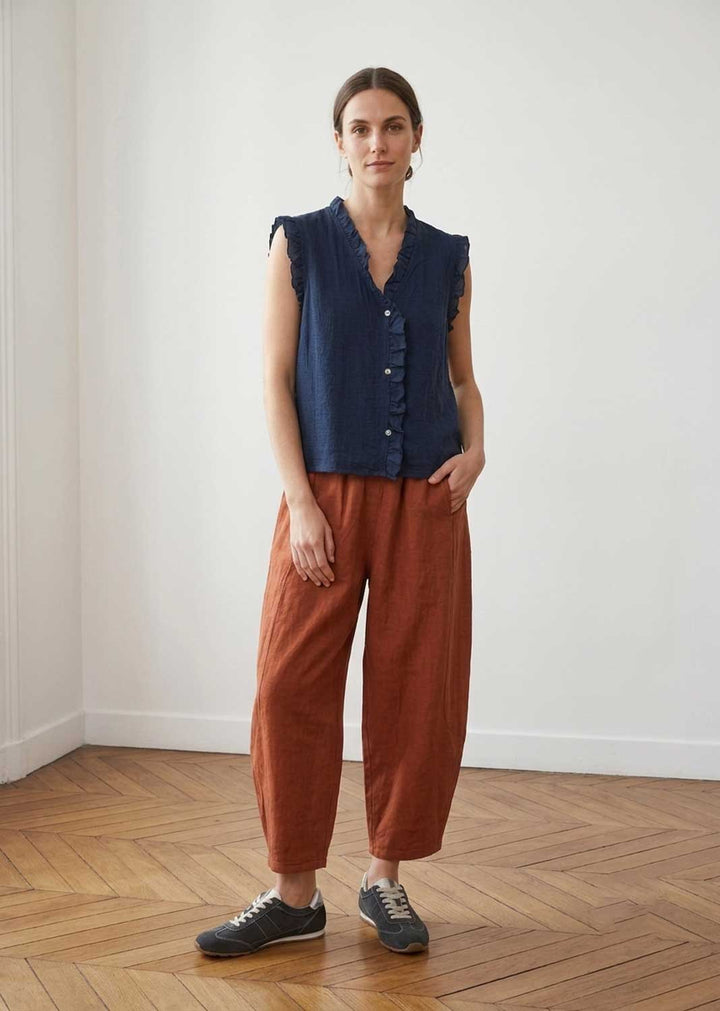A woman stands in a bright room with a wood floor and white walls, wearing a sleeveless navy blue button-front top and rust-brown wide-leg pants with dark gray sneakers.