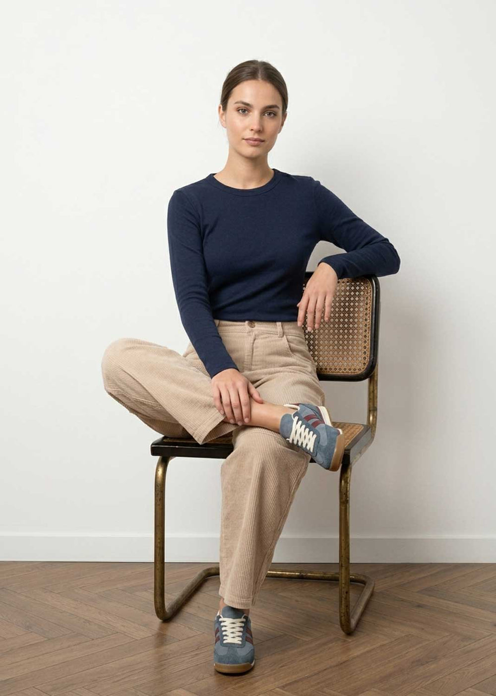 A woman sits on a metal and cane chair wearing a navy long-sleeve crewneck sweater, beige trousers, and blue sneakers against a white wall and wood floor.