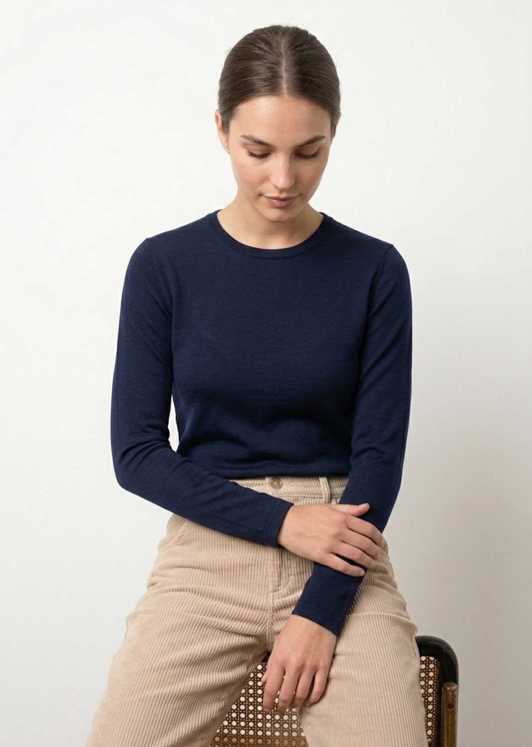 A woman wearing a navy blue long-sleeve crewneck sweater and beige corduroy trousers, seated on a chair against a plain light background.