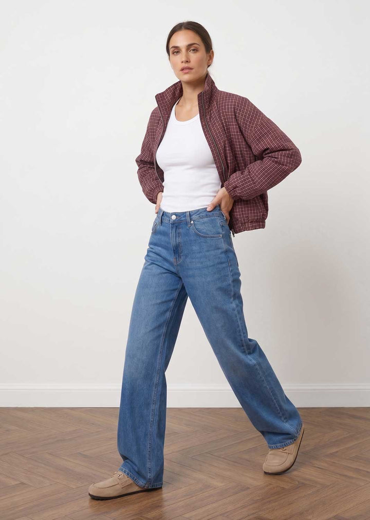 A woman modeling a pair of wide-leg blue jeans, styled with a white tank top, a maroon checkered zip-up jacket, and beige shoes, standing against a white wall with a wooden floor.