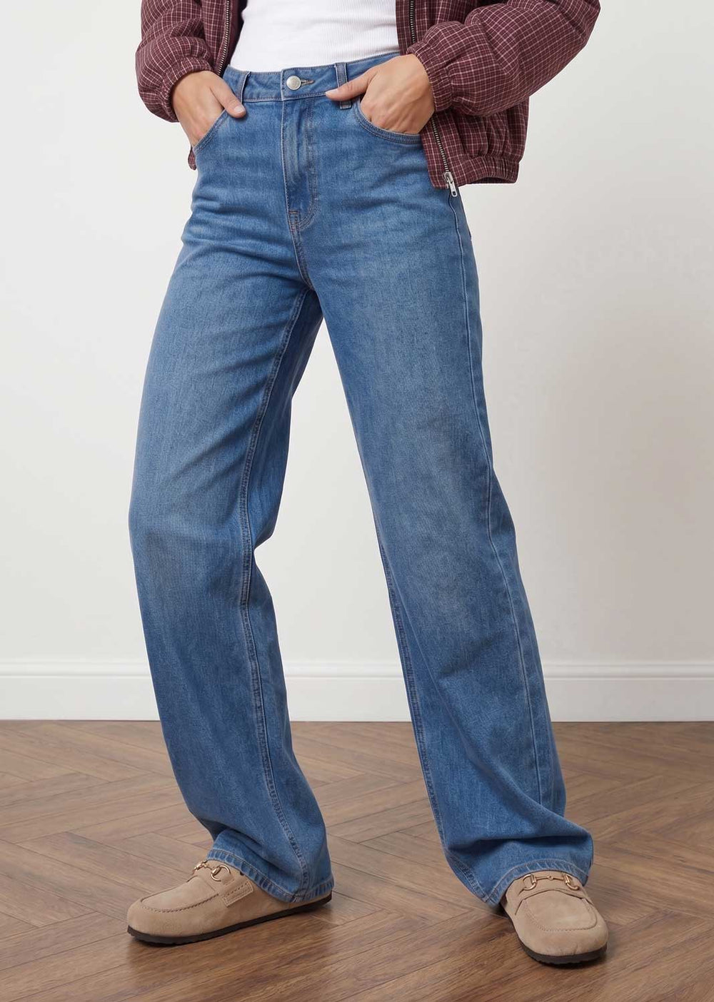 A person wearing blue wide-leg denim jeans, styled with a maroon plaid cardigan, white top, and beige suede shoes, standing on a wooden floor against a white wall.