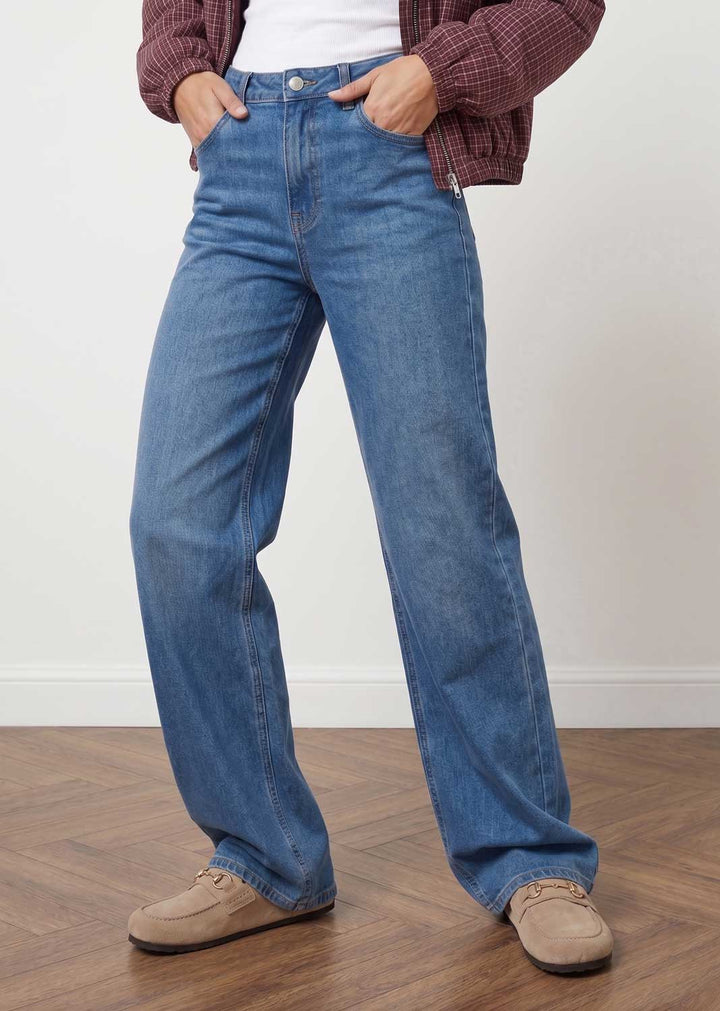 A person wearing blue wide-leg denim jeans, styled with a maroon plaid cardigan, white top, and beige suede shoes, standing on a wooden floor against a white wall.