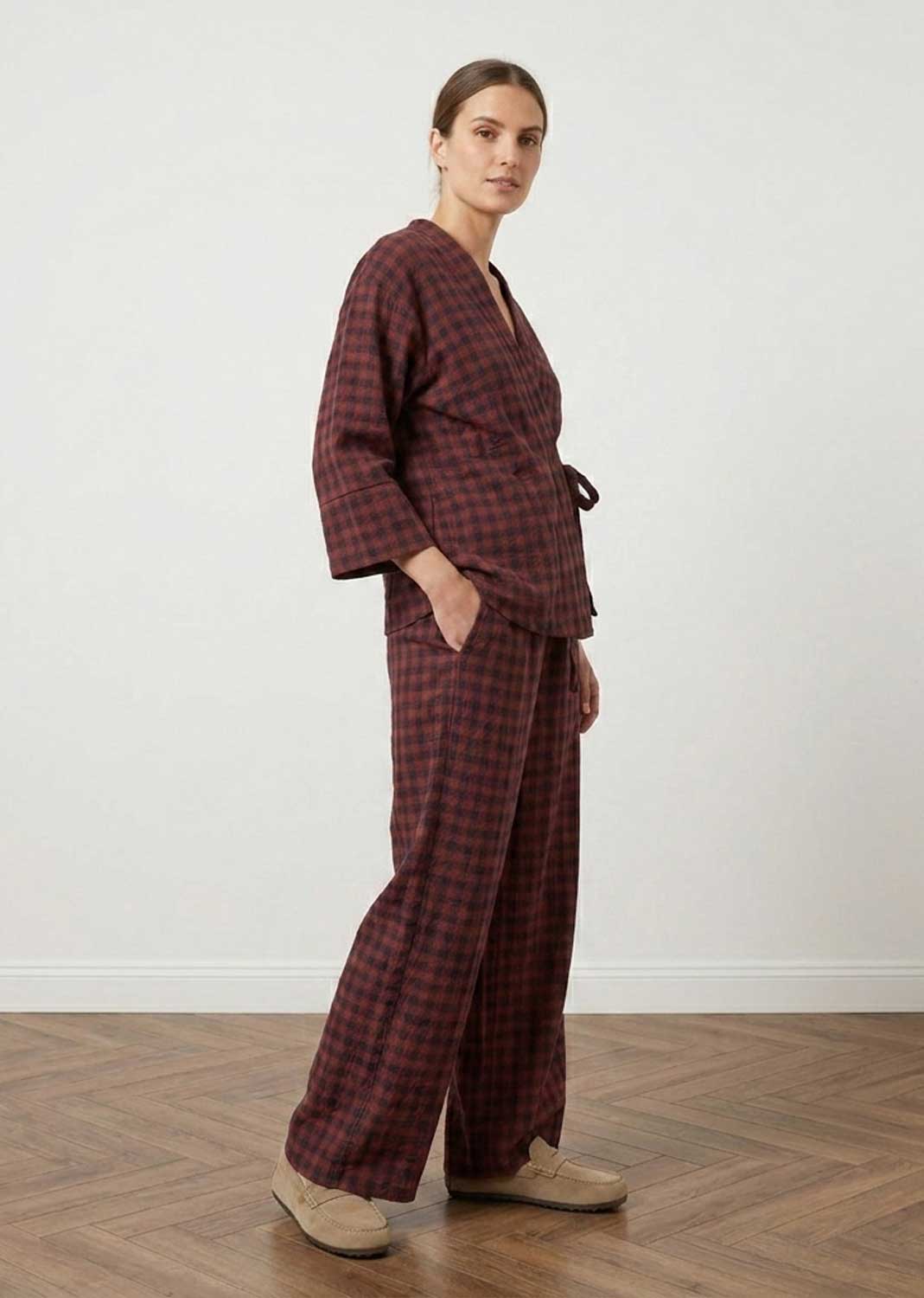 A woman modeling a matching two-piece plaid set in a dark red and black checkered pattern, standing in a minimalist room with a light gray wall and wooden parquet floor.