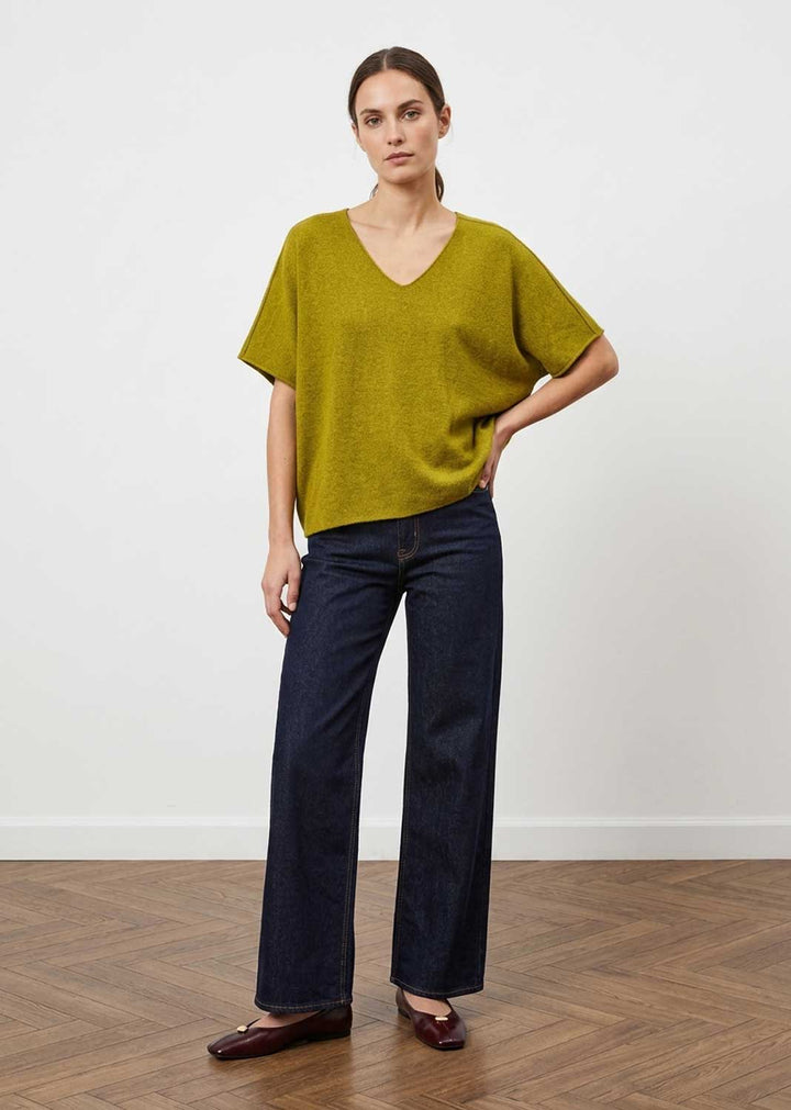 A woman modeling a short-sleeve V-neck knit top in olive yellow-green, paired with dark blue wide-leg jeans and dark brown flat shoes, standing against a white wall on a wood floor.