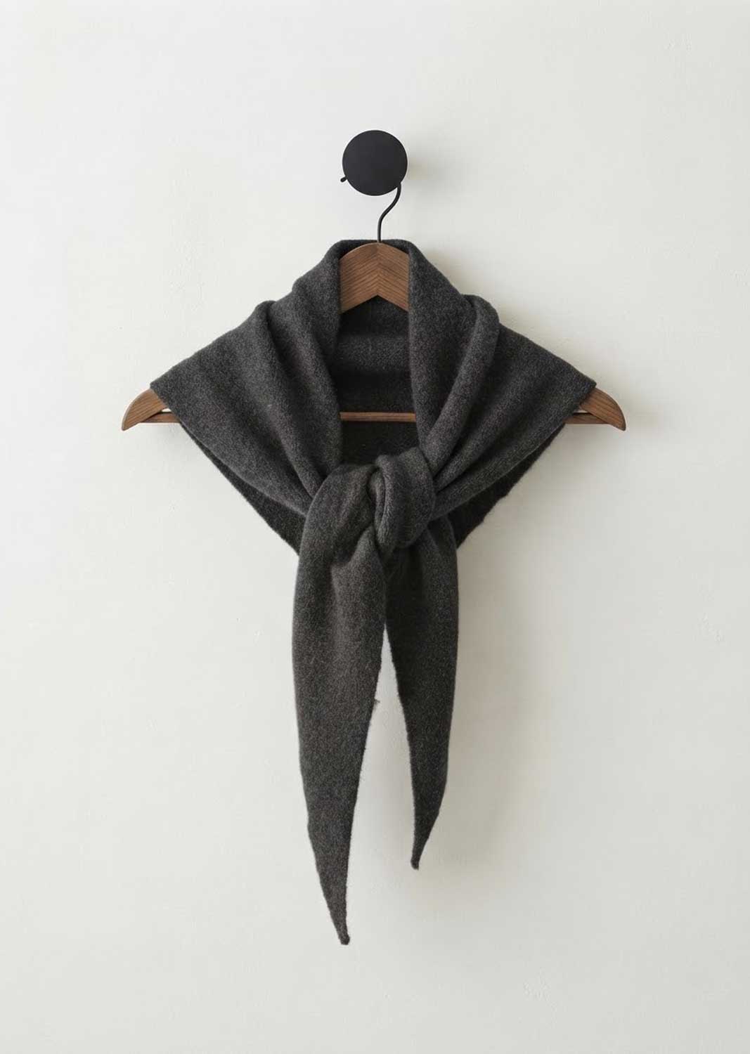 A dark charcoal gray knit scarf draped over a wooden hanger with a black wall hook above it, set against a plain off-white wall.