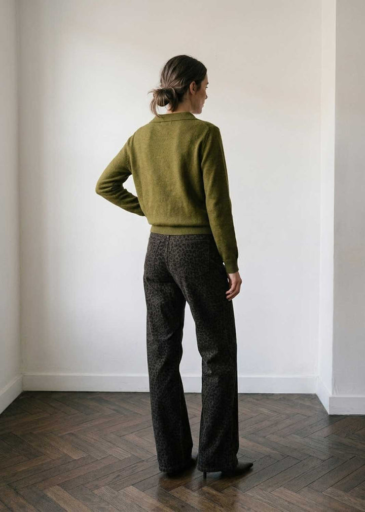A woman stands with her back to the camera in a minimalist room with white walls and a dark wood herringbone floor, wearing an olive green knit sweater and dark patterned trousers.