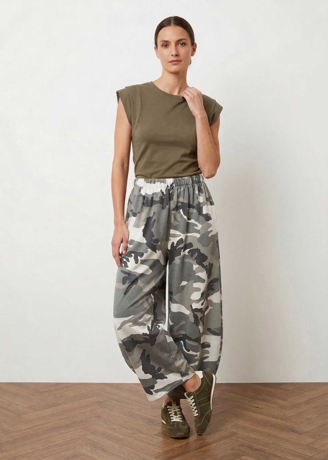 A woman modeling loose, baggy camouflage pants in shades of gray, white, and black, paired with an olive green sleeveless top and green sneakers, standing against a plain off-white wall and wooden floor.
