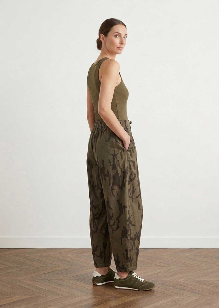 A woman stands in profile wearing an olive green sleeveless top and matching camouflage-print pants, paired with dark green sneakers, against a plain white wall and wood floor.
