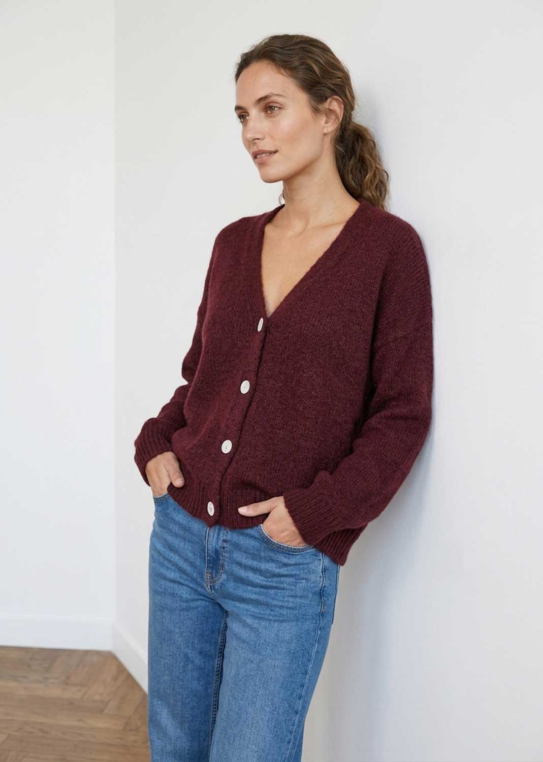 A woman wearing a maroon button-up wool sweater and blue jeans, standing against a white wall.